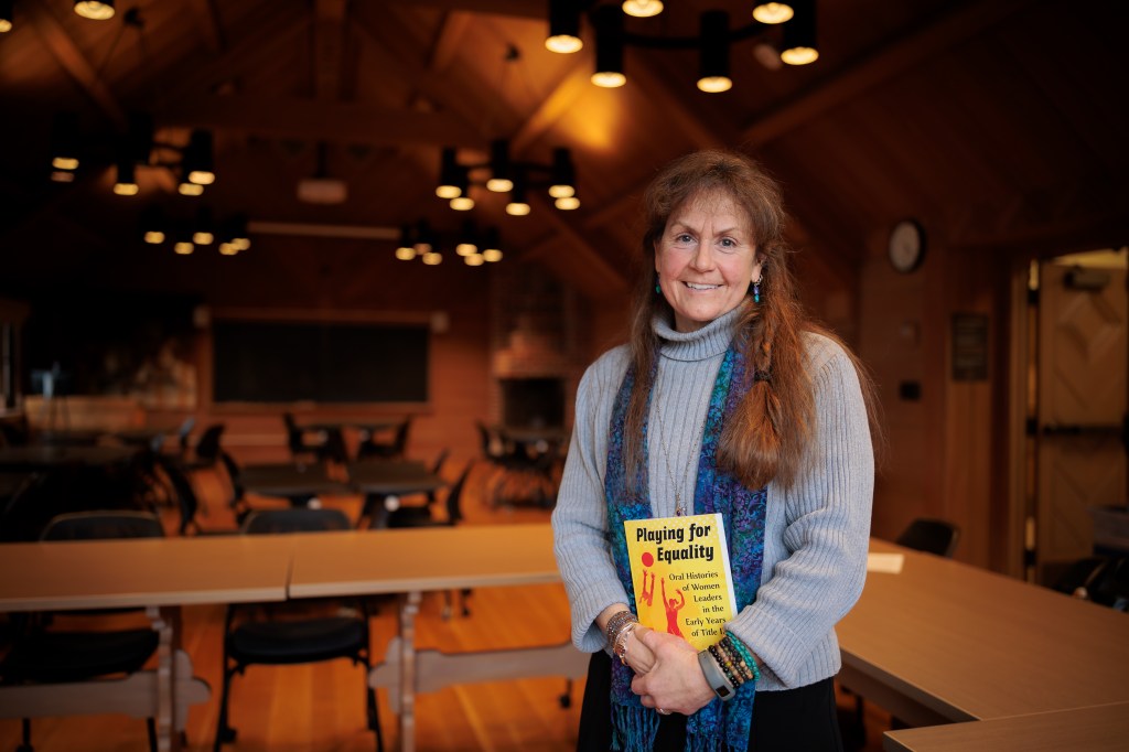 A professor poses for a photo with a book in a warmly-lit classroom.