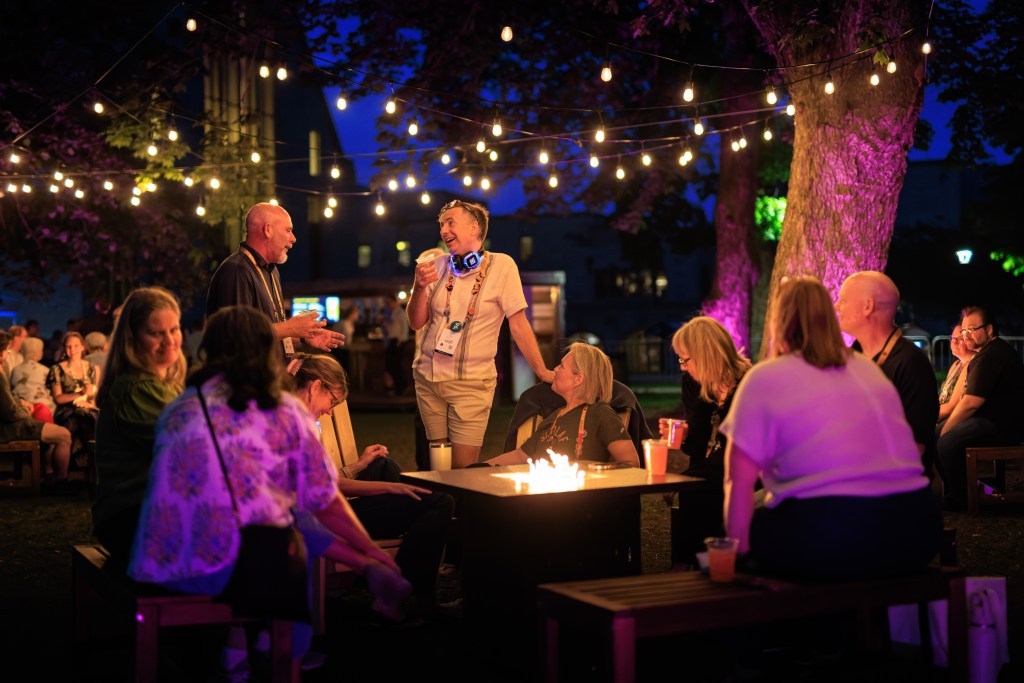 Alumni gather around a fire with string lights overhead.